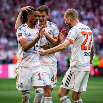 A Bayern München játékosai ünneplik egy csapattársuk gólját. Három játékos látható, akik örömmel beszélgetnek és gratulálnak egymásnak. A háttérben stadion és szurkolók láthatók, tükrözve a mérkőzés izgalmát.