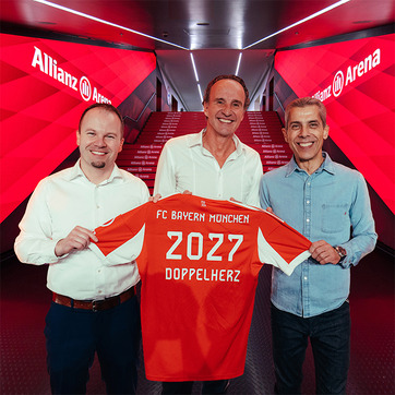 Három férfi áll egy színes háttér előtt, és bemutat egy piros focimezt. A mez hátán "FC Bayern München", "2027" és "DOPPELHERZ" feliratok láthatók. Az esemény az Allianz Arenában zajlik. Az arcuk boldog kifejezését mutatja az együttműködés örömére.